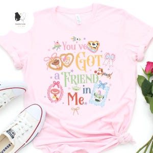 May include: A light pink t-shirt featuring the phrase "You've Got a Friend in Me" in colorful lettering. The shirt has various cartoon character illustrations and decorative frames. A pair of white sneakers are visible in the image.