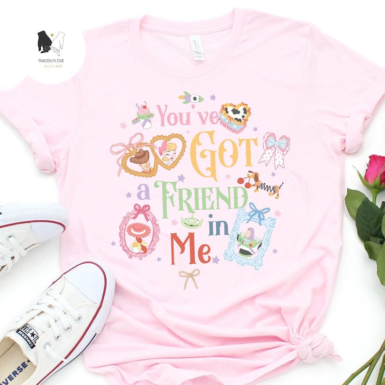 May include: A light pink t-shirt featuring the phrase "You've Got a Friend in Me" in colorful lettering. The shirt has various cartoon character illustrations and decorative frames. A pair of white sneakers are visible in the image.