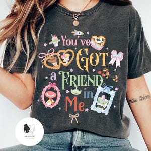 May include: Dark gray t-shirt with the phrase "You've Got a Friend in Me" in colorful text and framed cartoon characters. The shirt features a crew neck and short sleeves, and is worn with blue jeans.