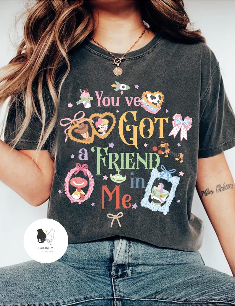 May include: Dark gray t-shirt with the phrase "You've Got a Friend in Me" in colorful text and framed cartoon characters. The shirt features a crew neck and short sleeves, and is worn with blue jeans.