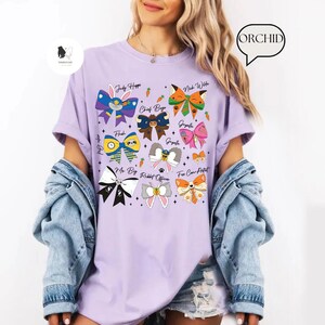 Puede incluir: Una camiseta lila con dise&ntilde;os de lazos de dibujos animados coloridos con nombres de personajes como Judy Hopps y Nick Wilde. La camiseta tiene mangas cortas y un corte holgado, ideal para el uso diario.