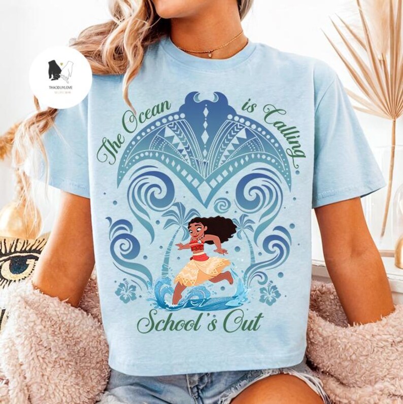 Op de afbeelding: Een lichtblauw t-shirt met een afbeelding van een personage met lang donker haar en een beige rok, omringd door blauwe golfpatronen, palmbomen en hibiscusbloemen. De tekst luidt The Ocean is Calling en School's Out.