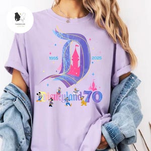 Op de afbeelding: Een paars T-shirt met een afbeelding van het Disneyland-kasteel en de tekst "Disneyland 70" met Mickey Mouse en zijn vrienden. De tekst "1955" en "2025" staan ook op het T-shirt.
