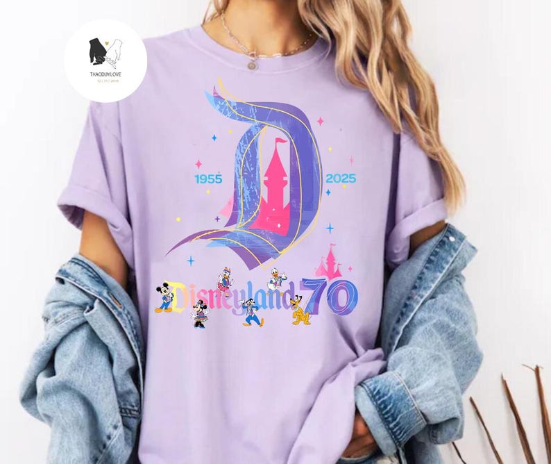 Op de afbeelding: Een paars T-shirt met een afbeelding van het Disneyland-kasteel en de tekst "Disneyland 70" met Mickey Mouse en zijn vrienden. De tekst "1955" en "2025" staan ook op het T-shirt.