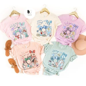 May include: Five pastel-colored t-shirts with Disney-themed graphics and text. Each shirt features a different design with characters and quotes. The shirts are displayed on gold hangers, with sandals and a hat nearby.