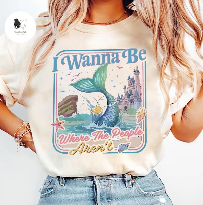 May include: Cream-colored t-shirt featuring a mermaid illustration with the text "I Wanna Be Where The People Aren't." The design includes a mermaid tail, castle, and ocean elements. The shirt has a vintage, slightly faded look.