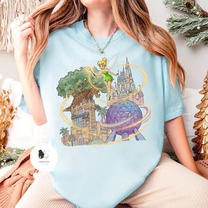 Op de afbeelding: Lichtblauw T-shirt met een kleurrijke afbeelding van Tinkerbell voor een boom, een kasteel en een geodetische bol. De afbeelding bevat de tekst "Hollywood Tower Hotel". Het shirt heeft een ronde hals en korte mouwen.