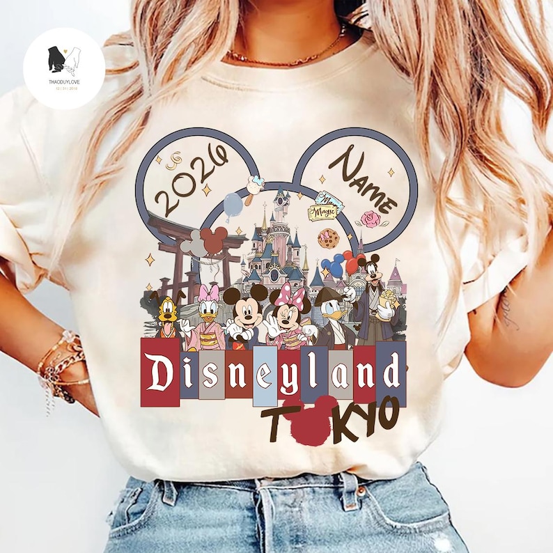 May include: Cream-colored t-shirt featuring a Disneyland Tokyo design. The design includes the text "2026" and "Name" within Mickey Mouse ears, along with various Disney characters and the words "Disneyland Tokyo".