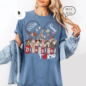 May include: Blue t-shirt featuring a Disneyland Tokyo design. The design includes Mickey Mouse ears with the year 2020 and a space for a name. The shirt showcases various Disney characters and the words "Disneyland Tokyo" in a red and white font.
