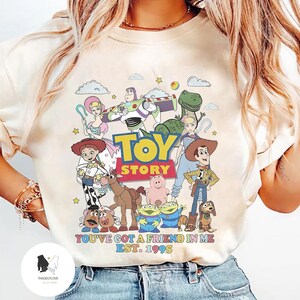 May include: A white t-shirt with a colorful graphic of characters from the Toy Story movie franchise. The graphic includes Woody, Buzz Lightyear, Jessie, Rex, Hamm, and Slinky Dog. The text "Toy Story" is printed in yellow and black letters. The text "You've got a friend in me Est. 1995" is printed in black letters below the graphic.