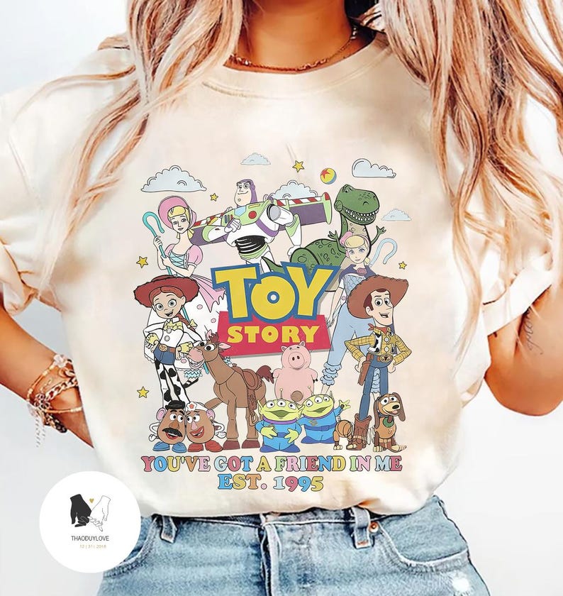 May include: A white t-shirt with a colorful graphic of characters from the Toy Story movie franchise. The graphic includes Woody, Buzz Lightyear, Jessie, Rex, Hamm, and Slinky Dog. The text "Toy Story" is printed in yellow and black letters. The text "You've got a friend in me Est. 1995" is printed in black letters below the graphic.