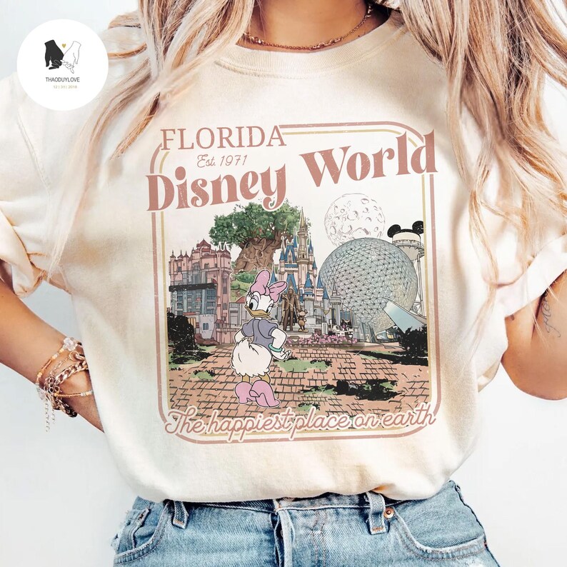 Puede incluir: Camiseta color crema con un gr&aacute;fico retro de Disney World en Florida. El dise&ntilde;o incluye las palabras "Florida Disney World Est. 1971" y un dibujo animado de Daisy Duck. Tambi&eacute;n se incluye la frase "The happiest place on earth".