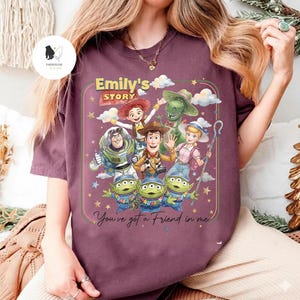 May include: A maroon t-shirt featuring a colorful graphic of characters from the animated film "Toy Story." The design includes Woody, Buzz Lightyear, Jessie, Rex, and the Little Green Men, with the text "Emily's Story" and "You've got a Friend in me."