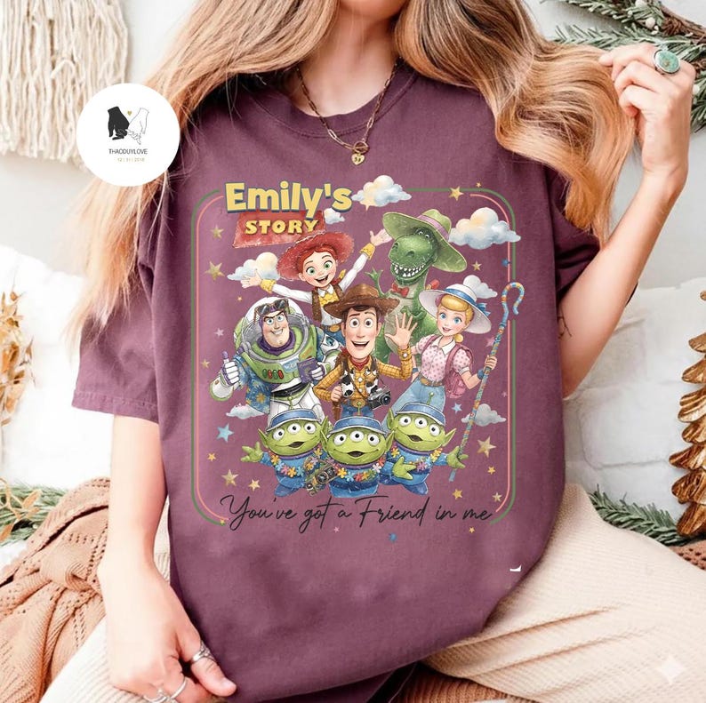 May include: A maroon t-shirt featuring a colorful graphic of characters from the animated film "Toy Story." The design includes Woody, Buzz Lightyear, Jessie, Rex, and the Little Green Men, with the text "Emily's Story" and "You've got a Friend in me."