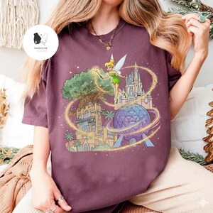 Op de afbeelding: Bordeauxrood T-shirt met een kleurrijke afbeelding van Tinkerbell, een boom, een kasteel en een geodetische bol. De tekst "Hollywood Tower Hotel" is zichtbaar. Het ontwerp is omgeven door gouden glitter.