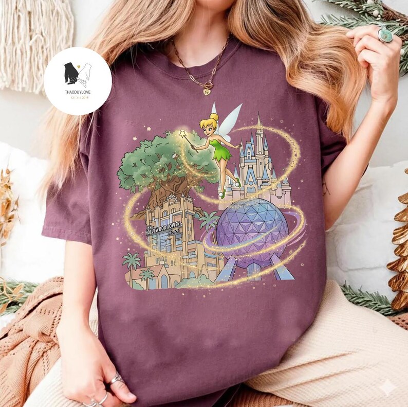 Op de afbeelding: Bordeauxrood T-shirt met een kleurrijke afbeelding van Tinkerbell, een boom, een kasteel en een geodetische bol. De tekst "Hollywood Tower Hotel" is zichtbaar. Het ontwerp is omgeven door gouden glitter.
