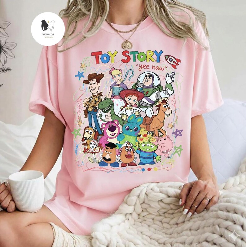 May include: A light pink t-shirt featuring a colorful graphic print of various Toy Story characters including Woody, Buzz Lightyear, Jessie, and others. The text "TOY STORY" and "yee haw" is printed above the characters with small star doodles.