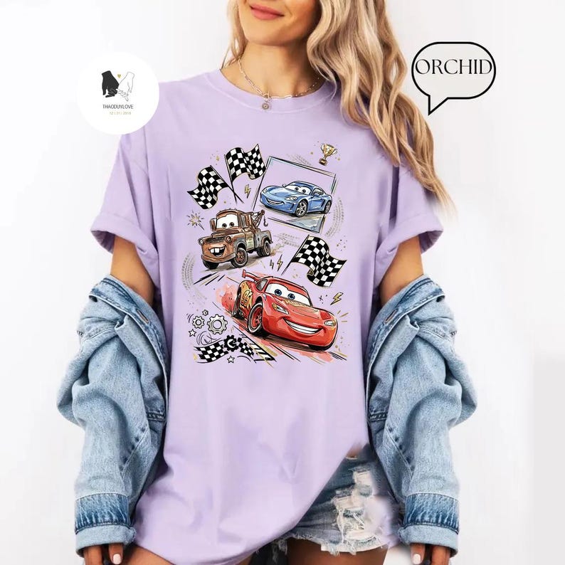 May include: A light purple t-shirt featuring a graphic of animated race cars, checkered flags, and a trophy. The design includes the characters Lightning McQueen, Mater, and other race cars. The shirt has short sleeves and a relaxed fit.