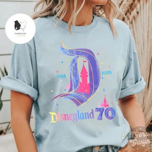 Op de afbeelding: Een lichtblauw T-shirt met een kleurrijke afbeelding van het Disneyland-kasteel en de tekst "Disneyland 70" met de jaren 1955 en 2025.