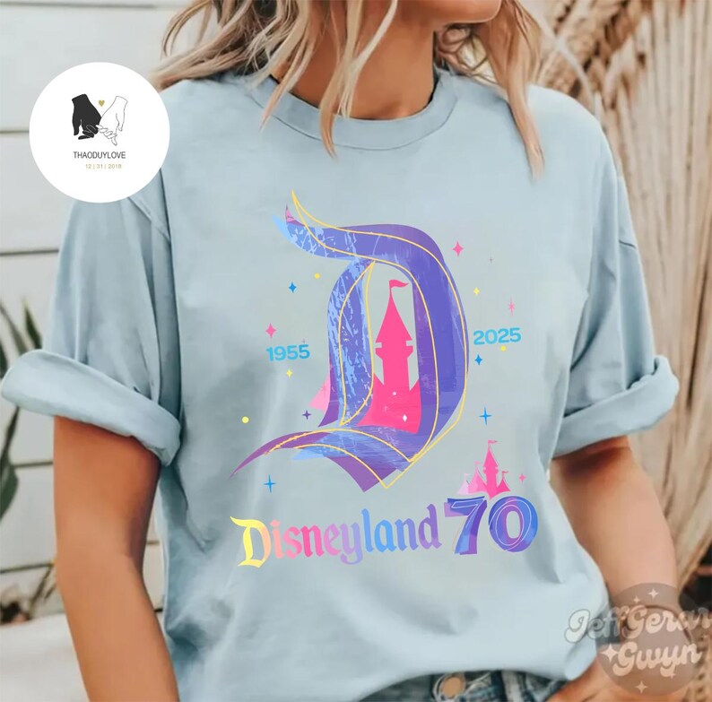 Op de afbeelding: Een lichtblauw T-shirt met een kleurrijke afbeelding van het Disneyland-kasteel en de tekst "Disneyland 70" met de jaren 1955 en 2025.