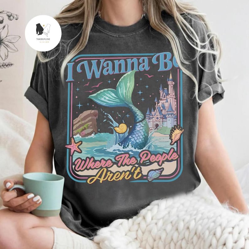 May include: Dark gray t-shirt with a mermaid graphic. The design features a mermaid with a teal tail, a castle, and the text "I Wanna Be Where The People Aren't" in a retro style. A starfish and seashells are also included.