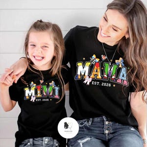 May include: Two black t-shirts with colourful text and butterfly accents. One shirt says "MIMI EST. 2026" and the other says "MAMA EST. 2026". The shirts are worn by a mother and daughter.