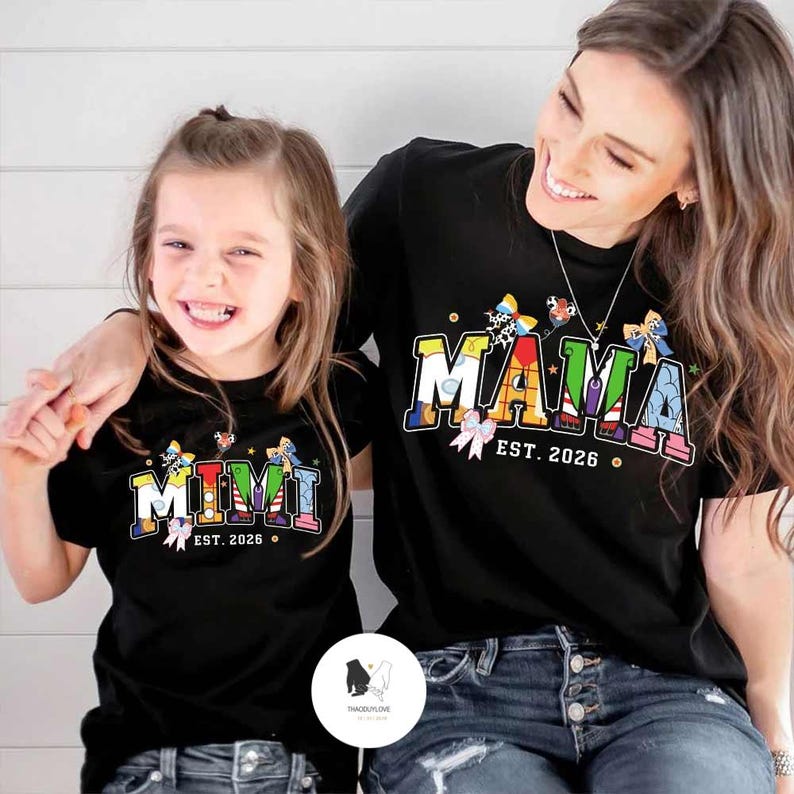 May include: Two black t-shirts with colourful text and butterfly accents. One shirt says "MIMI EST. 2026" and the other says "MAMA EST. 2026". The shirts are worn by a mother and daughter.