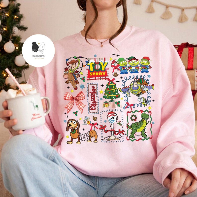 May include: A light pink sweatshirt featuring a colorful Toy Story graphic. The design includes images of Woody, Buzz Lightyear, the Little Green Men, and other characters, along with Christmas-themed elements. The sweatshirt has long sleeves.