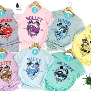 May include: Seven different t-shirts featuring characters from the Disney Pixar movie Cars. Each shirt has a different character, including McQueen, Mater, Lightning McQueen, Sally, Guido, Luigi, and Doc Hudson. The shirts are all in different colours, including grey, pink, blue, yellow, and green.