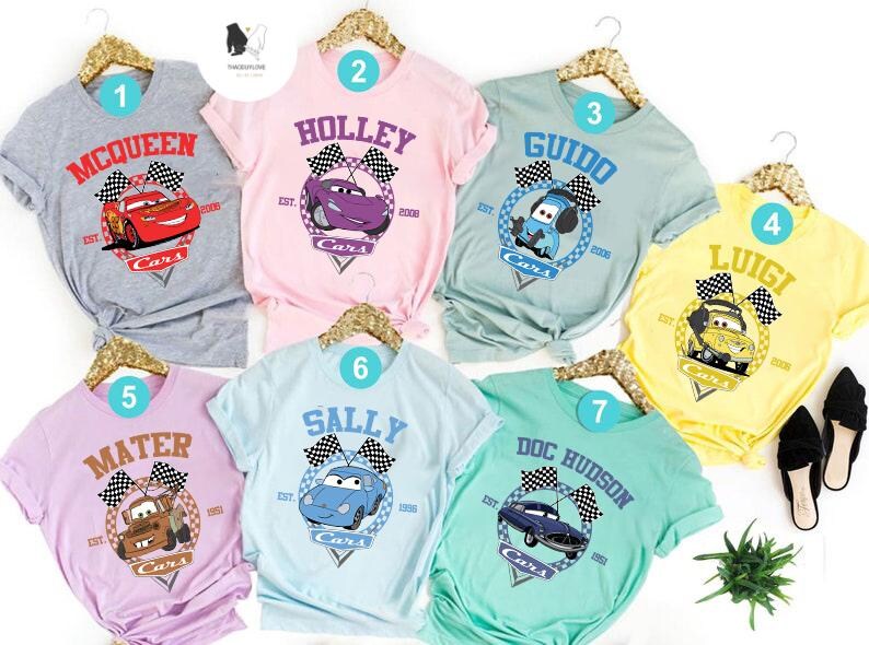 May include: Seven different t-shirts featuring characters from the Disney Pixar movie Cars. Each shirt has a different character, including McQueen, Mater, Lightning McQueen, Sally, Guido, Luigi, and Doc Hudson. The shirts are all in different colours, including grey, pink, blue, yellow, and green.