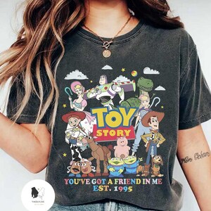 May include: A dark gray t-shirt with a graphic print of characters from the Toy Story movie franchise. The graphic includes Woody, Buzz Lightyear, Jessie, Rex, Hamm, Slinky Dog, and other characters. The text "Toy Story" is printed in white letters above the characters. The text "You've got a friend in me Est. 1995" is printed in white letters below the characters.