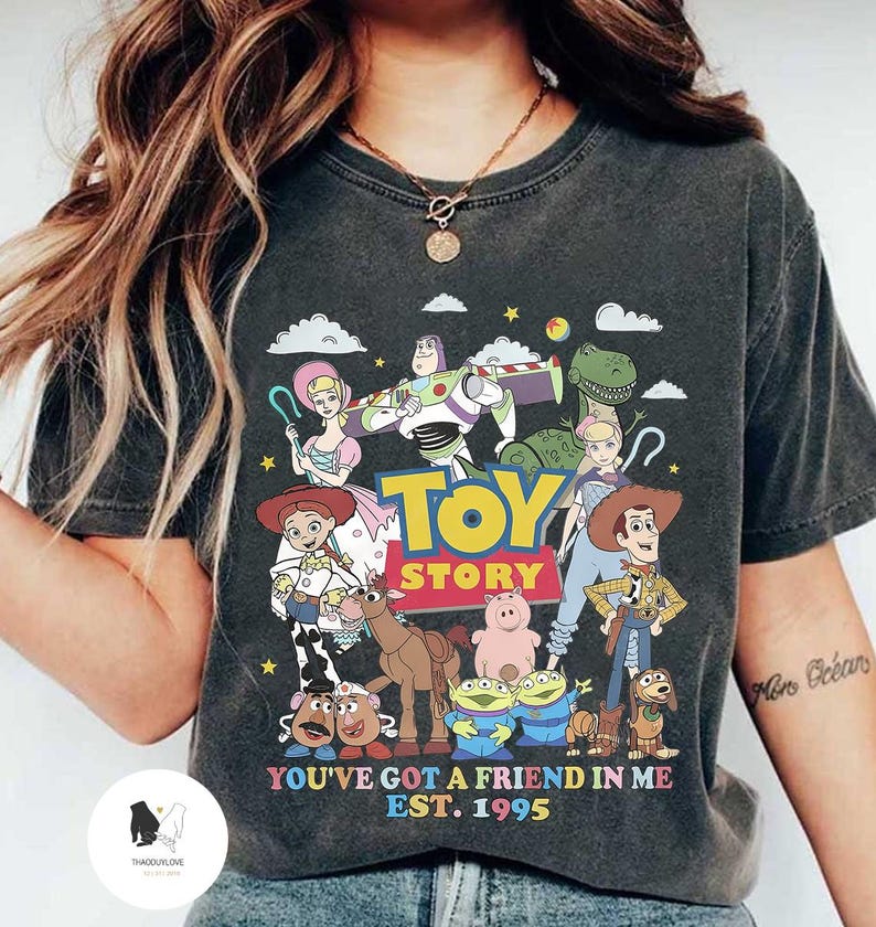 May include: A dark gray t-shirt with a graphic print of characters from the Toy Story movie franchise. The graphic includes Woody, Buzz Lightyear, Jessie, Rex, Hamm, Slinky Dog, and other characters. The text "Toy Story" is printed in white letters above the characters. The text "You've got a friend in me Est. 1995" is printed in white letters below the characters.