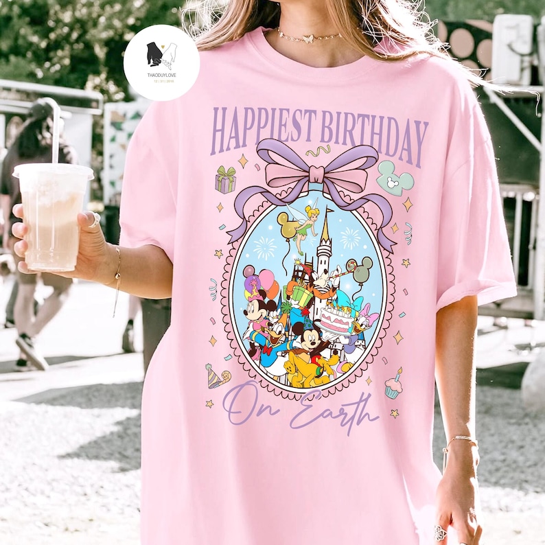 May include: Pink t-shirt with the text "Happiest Birthday On Earth" in purple script. The shirt features a colorful graphic of Disney characters, including Mickey and Minnie Mouse, in an oval frame with a bow.