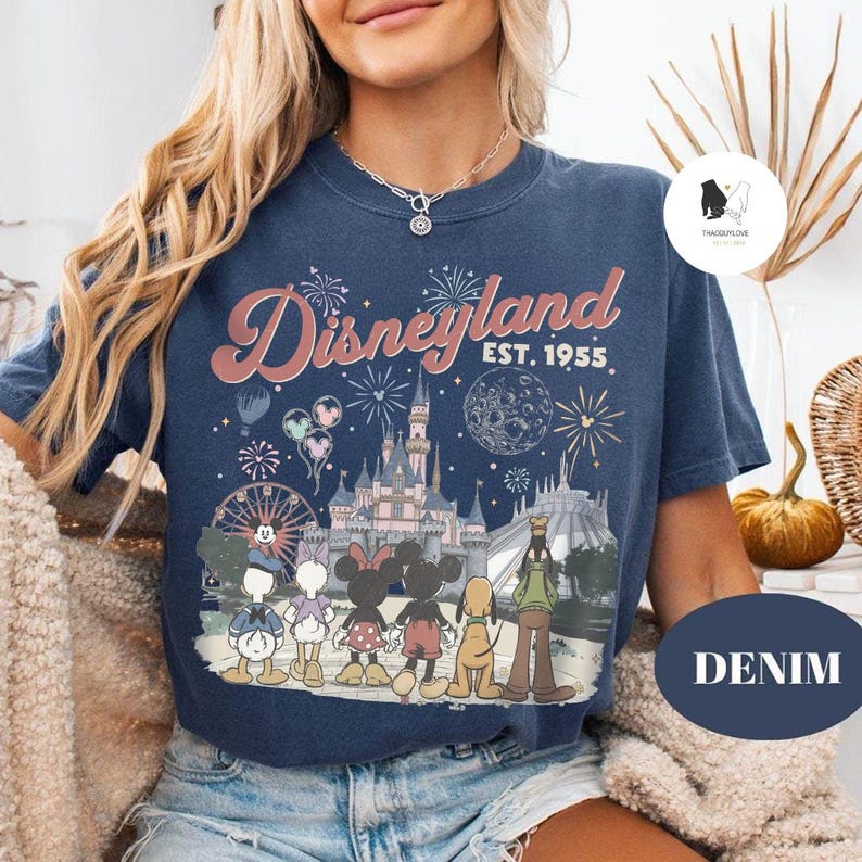 May include: A navy blue t-shirt featuring a graphic print of Disneyland with the text Disneyland Est. 1955. The design includes a castle, fireworks, a moon, and cartoon characters including Donald Duck, Daisy Duck, Minnie Mouse, Mickey Mouse, Pluto, and Goofy.