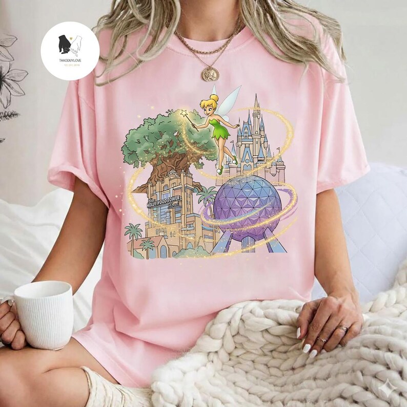 Op de afbeelding: Lichtroze t-shirt met een kleurrijke afbeelding van Tinkerbell, een boom, een kasteel en een grote bol. De afbeelding bevat de tekst "Hollywood Tower Hotel". Het shirt is gemaakt van een zacht materiaal.
