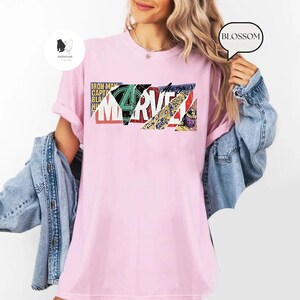 Op de afbeelding: Lichtroze t-shirt met een kleurrijk Marvel-logo. De afbeelding bevat het woord "Marvel" in vette letters, samen met andere tekst en stripboekachtige afbeeldingen. Een casual kledingstuk voor dagelijks gebruik.