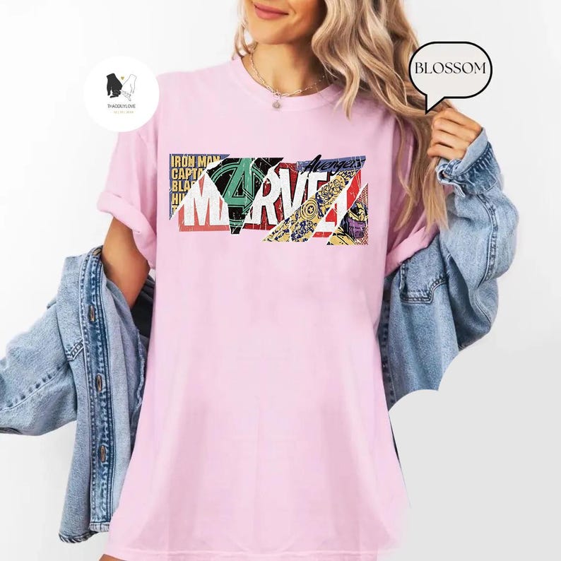 Op de afbeelding: Lichtroze t-shirt met een kleurrijk Marvel-logo. De afbeelding bevat het woord "Marvel" in vette letters, samen met andere tekst en stripboekachtige afbeeldingen. Een casual kledingstuk voor dagelijks gebruik.