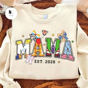 May include: Cream-coloured sweatshirt featuring the word "MAMA" in colourful, cartoon-style lettering. The design includes bow accents and the text "EST. 2026". A fun, playful design for a new mother.