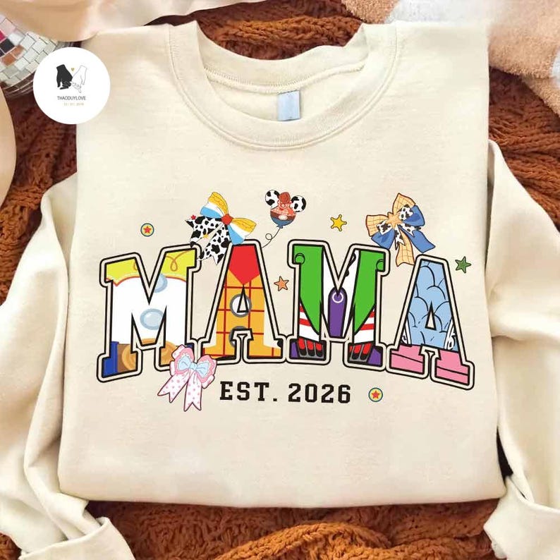 May include: Cream-coloured sweatshirt featuring the word "MAMA" in colourful, cartoon-style lettering. The design includes bow accents and the text "EST. 2026". A fun, playful design for a new mother.