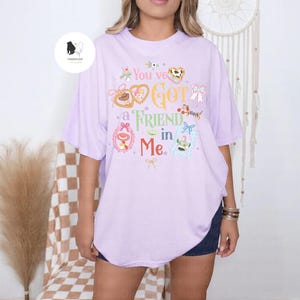 May include: A light purple t-shirt with the phrase "You've Got a Friend in Me" in colorful text and illustrations. The design includes hearts, bows, and character images. The shirt is a relaxed fit.
