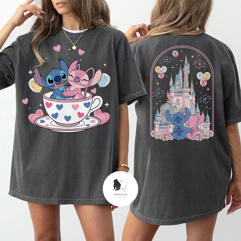 May include: Dark gray t-shirt featuring a cartoon illustration of Stitch and Angel in a teacup with hearts. The back of the shirt shows a castle with Stitch and Angel. The shirt has a crew neck and short sleeves.