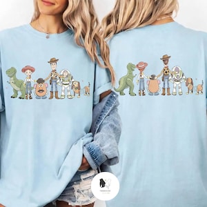 Peut inclure: T-shirts bleu clair avec une illustration de dessin anim&eacute; d'un groupe de personnages. Les personnages comprennent un dinosaure vert, une cow-girl, un cow-boy, une t&ecirc;te de pomme de terre, un astronaute, un chien et un chien avec une queue en ressort. Les chemises ont un col rond.