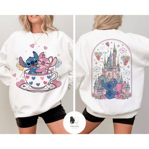 May include: White sweatshirt featuring a cartoon illustration of Stitch and Angel hugging in a teacup with hearts. The back of the sweatshirt shows Stitch and Angel in front of a castle with fireworks and balloons. The brand name is at the bottom.