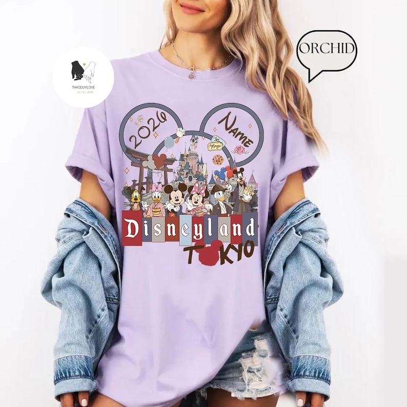 May include: A light purple t-shirt with a Disneyland Tokyo graphic. The graphic features Mickey Mouse ears with the text "2026" and "Name". Below the ears is a colorful illustration of Disney characters and landmarks. The words "Disneyland Tokyo" are printed in a bold font.