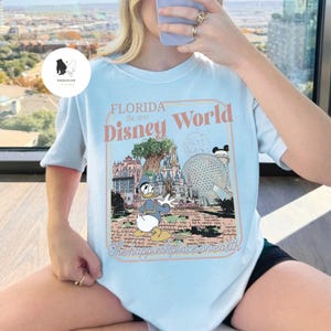 Puede incluir: Camiseta azul claro con un dise&ntilde;o de Donald Duck frente a los monumentos de Disney World. El dise&ntilde;o incluye el texto "FLORIDA Disney World" y "The happiest place on earth" en un marco color melocot&oacute;n.