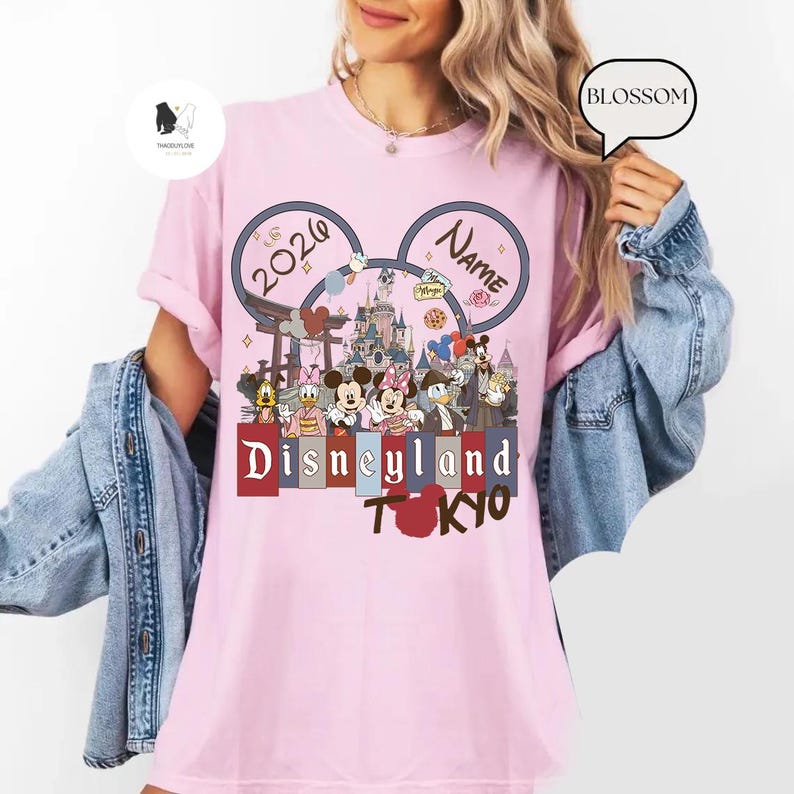 May include: Pink t-shirt featuring a Disneyland Tokyo design with Mickey and Minnie Mouse, and other Disney characters. The design includes the text "2026" and "Name". The shirt has short sleeves.