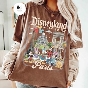 May include: Brown t-shirt featuring a Disneyland Paris graphic with Mickey Mouse, Donald Duck, and other Disney characters. The design includes the Eiffel Tower and Arc de Triomphe. The text "Disneyland Est. 1992" is also visible.