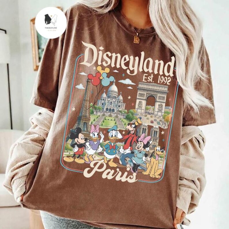 May include: Brown t-shirt featuring a Disneyland Paris graphic with Mickey Mouse, Donald Duck, and other Disney characters. The design includes the Eiffel Tower and Arc de Triomphe. The text "Disneyland Est. 1992" is also visible.