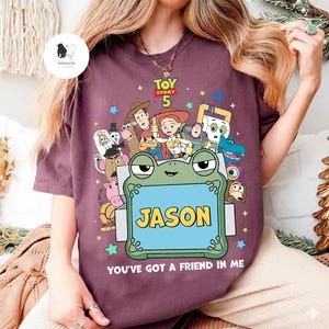 May include: A maroon t-shirt featuring a colorful graphic of characters from the movie Toy Story 5. The design includes a frog holding a sign that says "JASON" and the phrase "YOU'VE GOT A FRIEND IN ME".