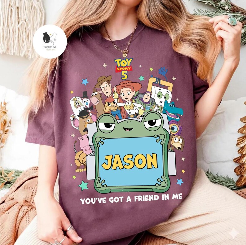 May include: A maroon t-shirt featuring a colorful graphic of characters from the movie Toy Story 5. The design includes a frog holding a sign that says "JASON" and the phrase "YOU'VE GOT A FRIEND IN ME".
